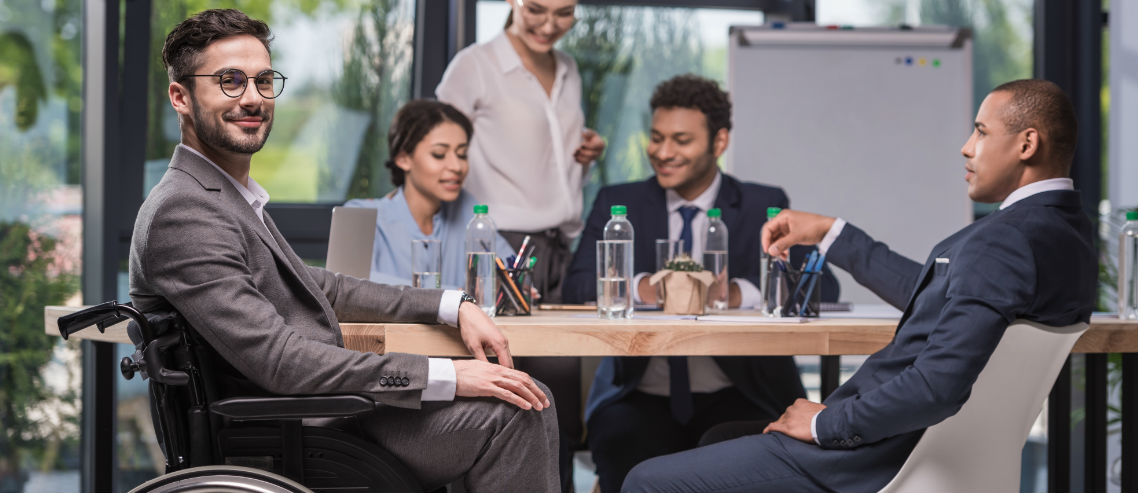 a man in a wheelchair sits in the foreground whilst his professional work colleagues discuss something in the background, symbolizing the importance of understanding how diversity and inclusion can be supported by your group insurance and employee benefits