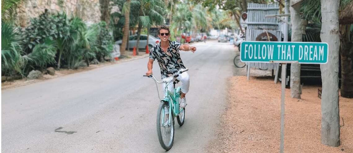 Man smiling while riding bicycle on paved road with follow your dream sign