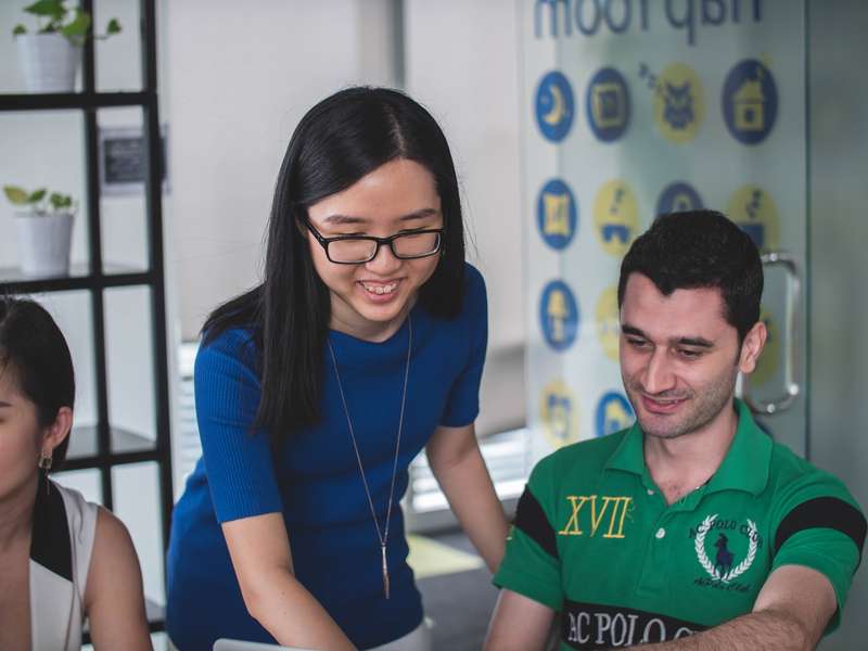 In 2023, A Smiling Woman Stands Beside A Smiling Man, Pointing At A Macbook, Illustrating Employee Benefits Plans