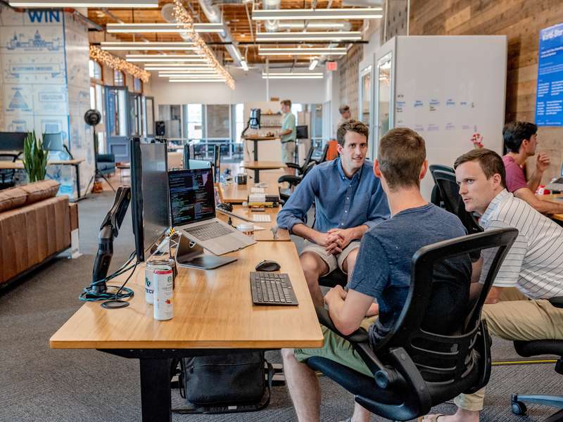 Three Men Sitting On Chairs Beside Tables, Discussing How To Design Employee Benefits Plans In 2023.